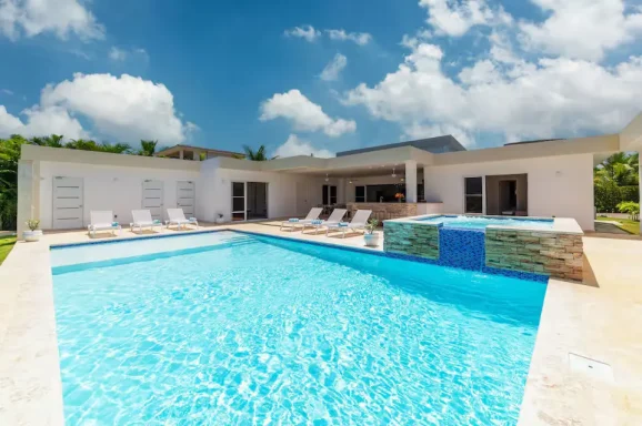 Moderne Villa mit Pool und Liegen, umgeben von Grün und blauen Himmel.