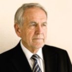 Ein seriöser Mann in Anzug mit gestreifter Krawatte vor neutralem Hintergrund.